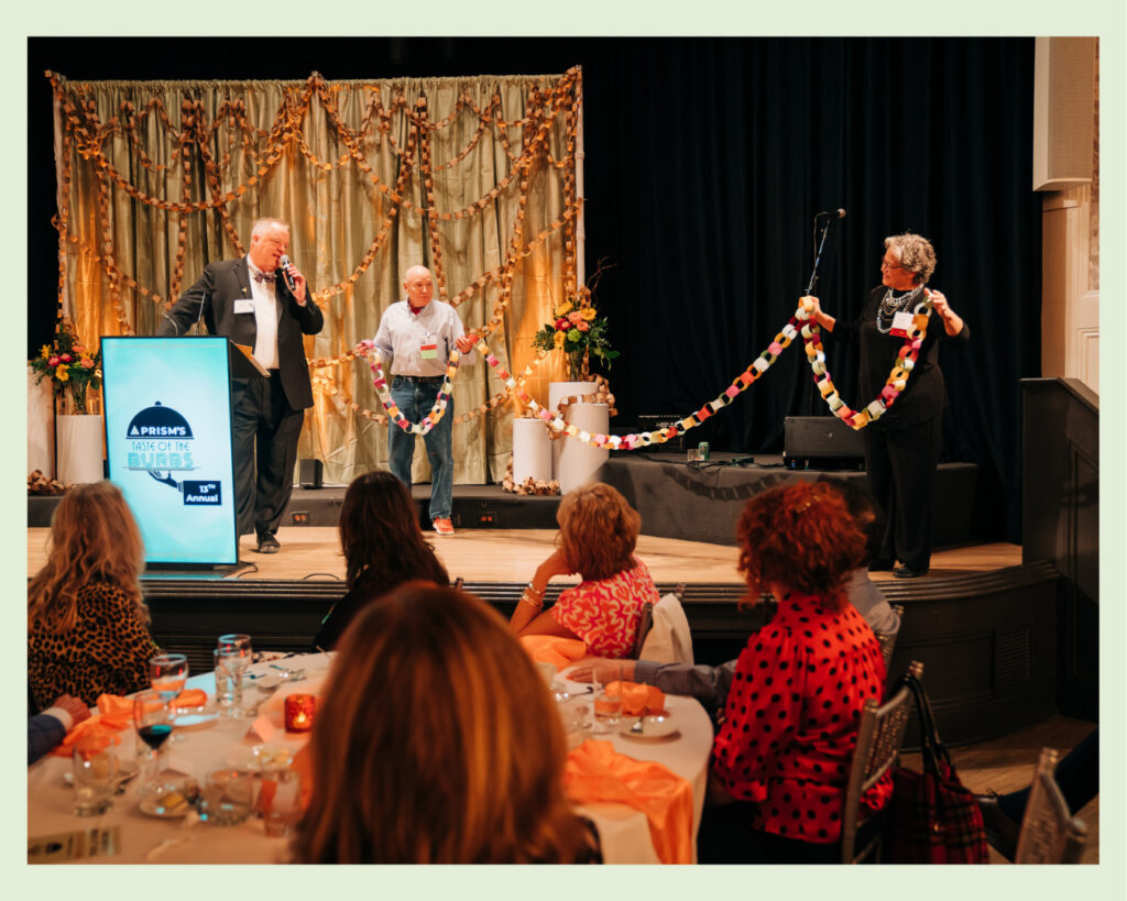 Man on stage holds one end of a paper chain and a woman holds the other end of a paper chain. Another man is standing to the left of the first man and holding a microphone and speaking into it.