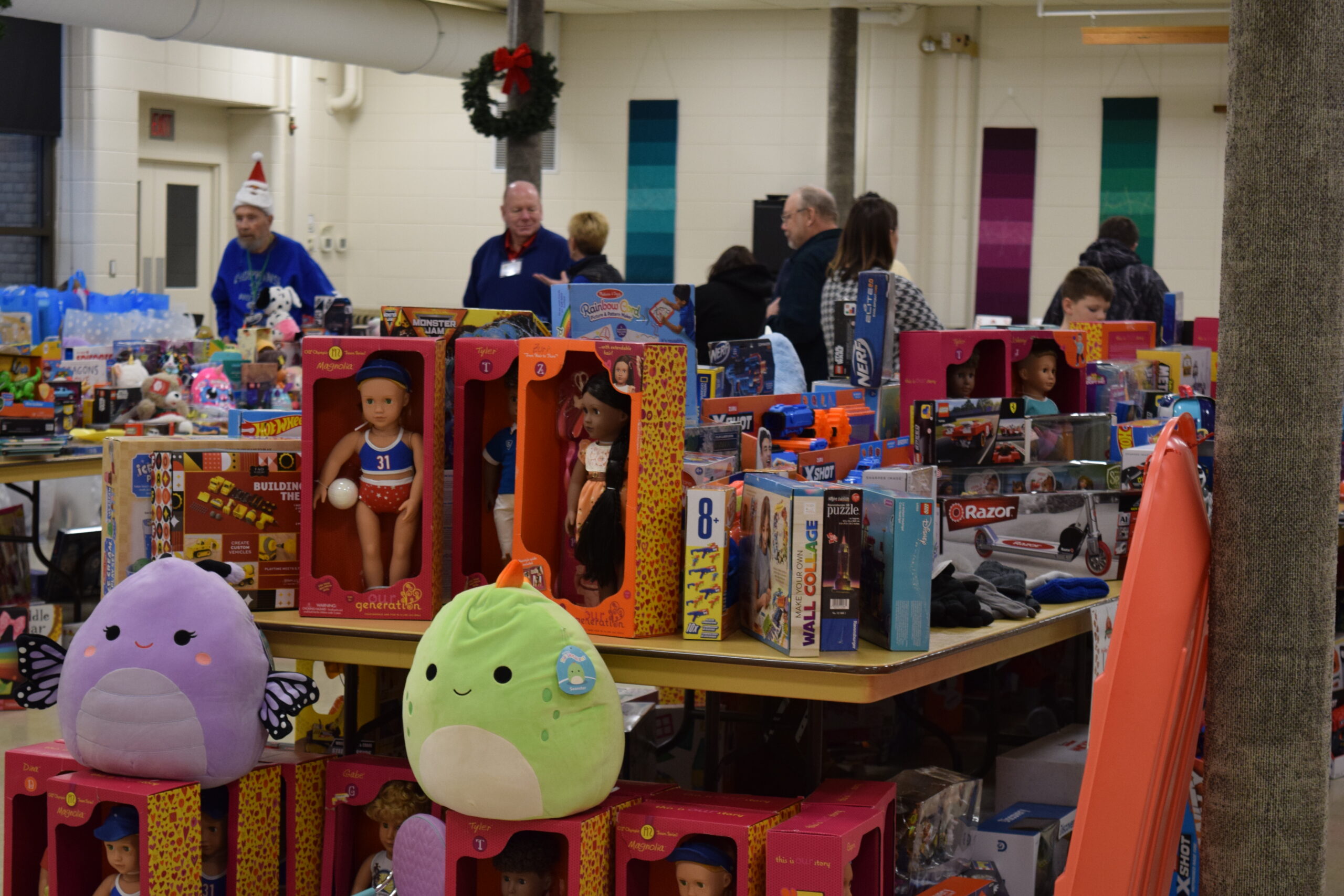 A table set up like a display filled with toys, including dolls, stuffed animals, building kits. There are volunteers in the background putting out toys onto the table.